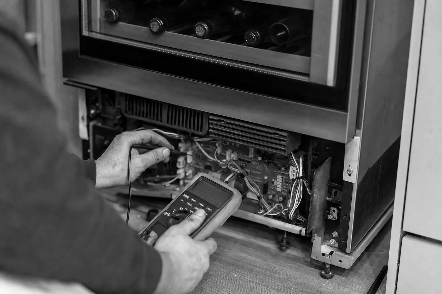 Wine cooler with its lower panel off and a meter probe on the exposed control board below the bottle racks