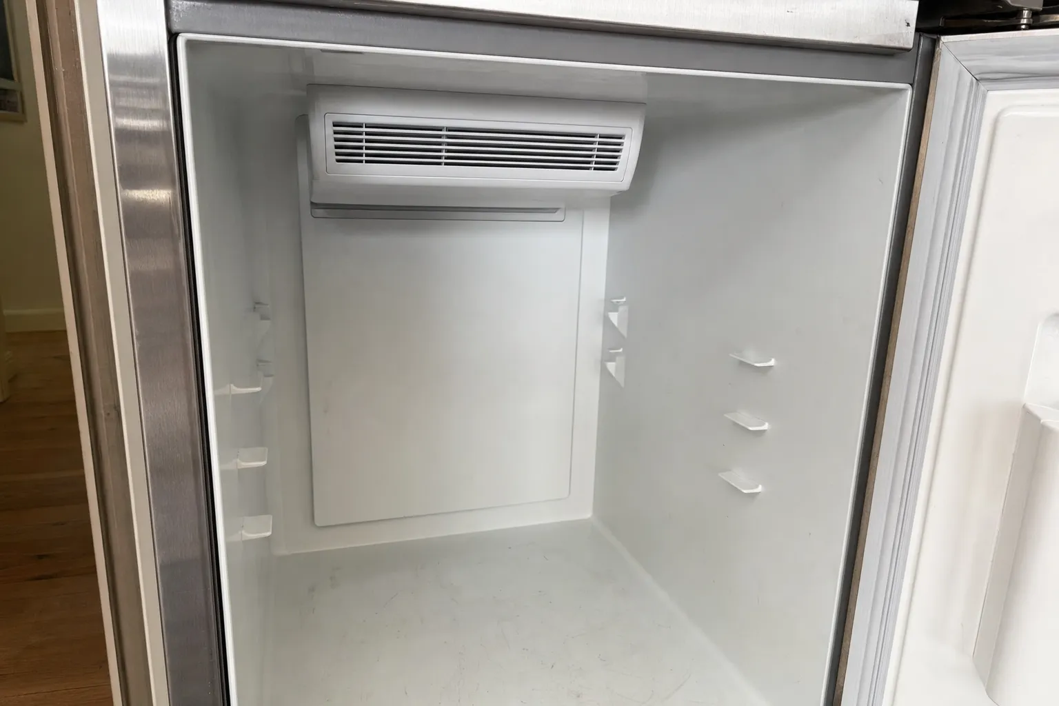 Empty white interior of a stainless refrigerator compartment, door open, the air vent cover at the top and shelf supports on both walls